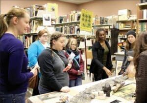 A group of people standing around a table in a library, looking at objects displayed on the surface.