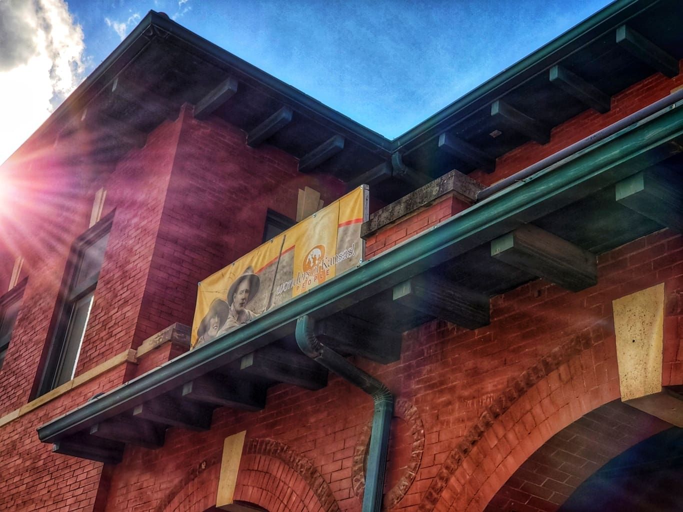 Sunlight flares over the corner of a historic red brick building with a dark green roof and a mural on the exterior.