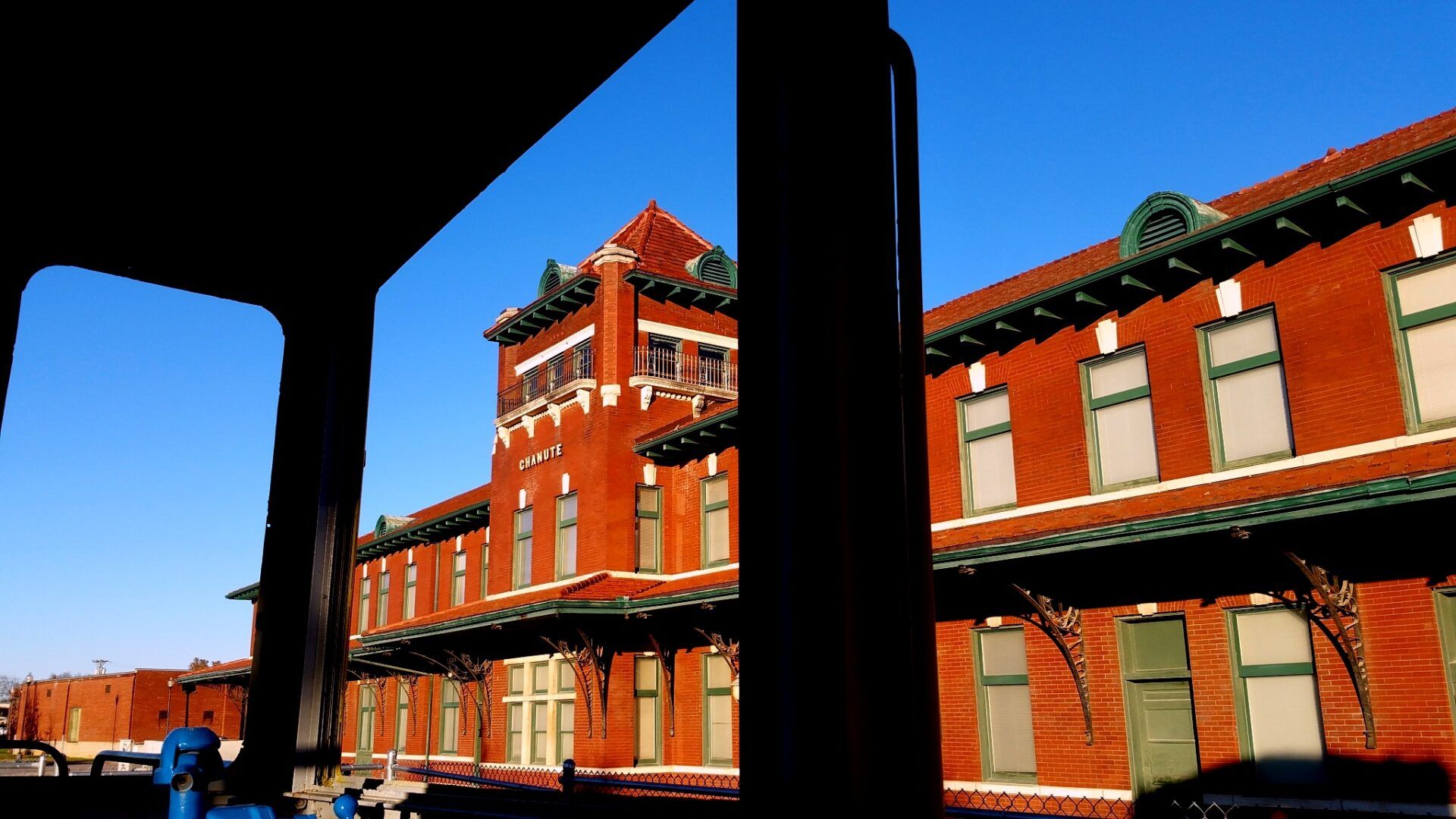 A red brick train depot with a central tower viewed through a dark window frame under a clear blue sky.