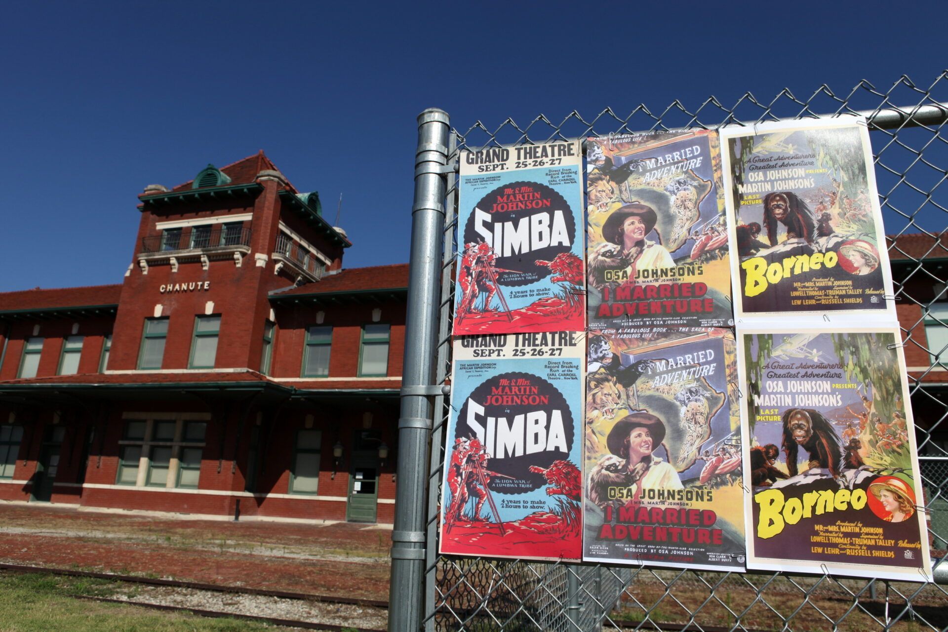 Posters for the films Simba and Borneo are attached to a chain-link fence in front of a red brick historic depot.