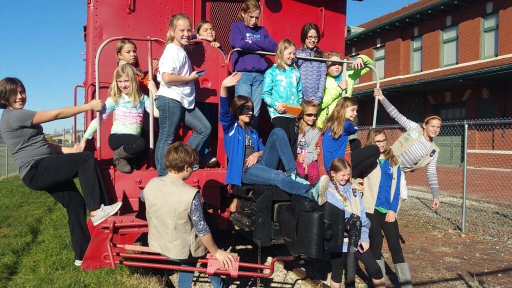 A group of people poses on and around a red train car outdoors near a brick building.