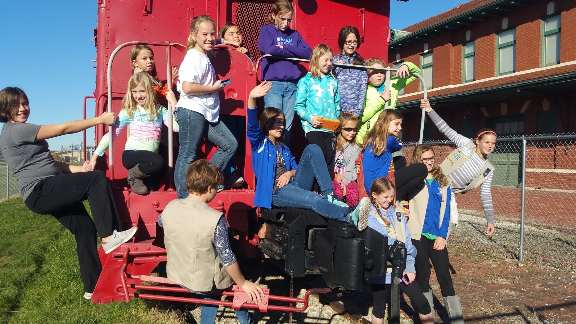 A group of people poses on and around a large red train car at a train depot on a sunny day.