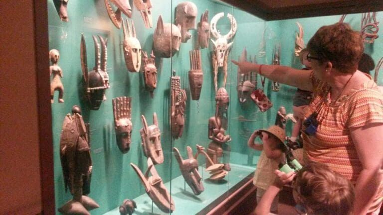 A visitor points to a display of wooden African masks inside a glass case at a museum.