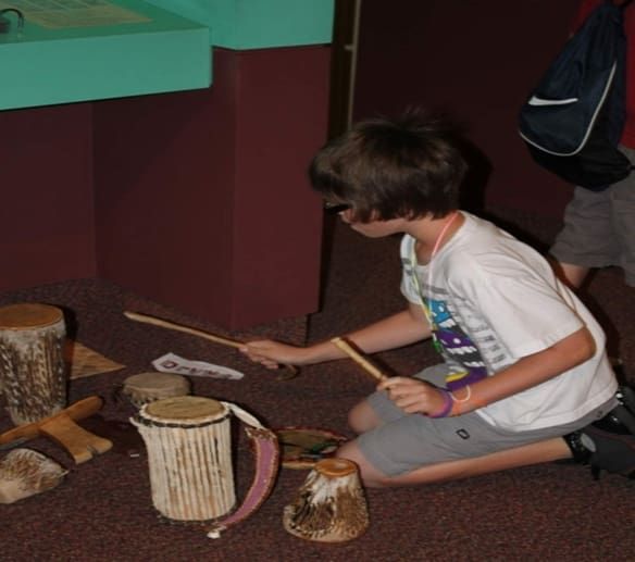 A person kneeling on a carpeted floor, playing wooden drums with sticks in an educational or museum exhibit.