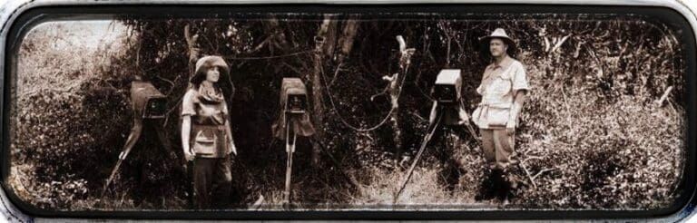 Two people standing in a forest, each beside a camera on a tripod, in a vintage sepia-toned photograph.