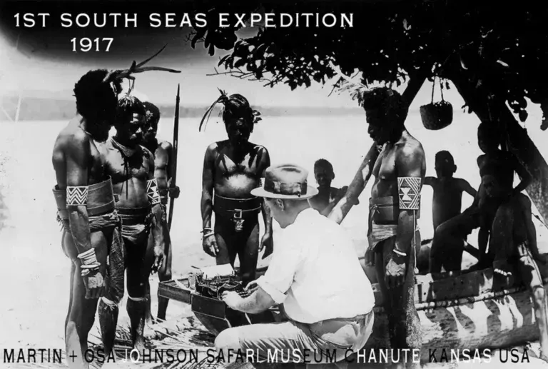 A man in a pith helmet and shirt interacts with indigenous people near a canoe at the 1917 South Seas Expedition.