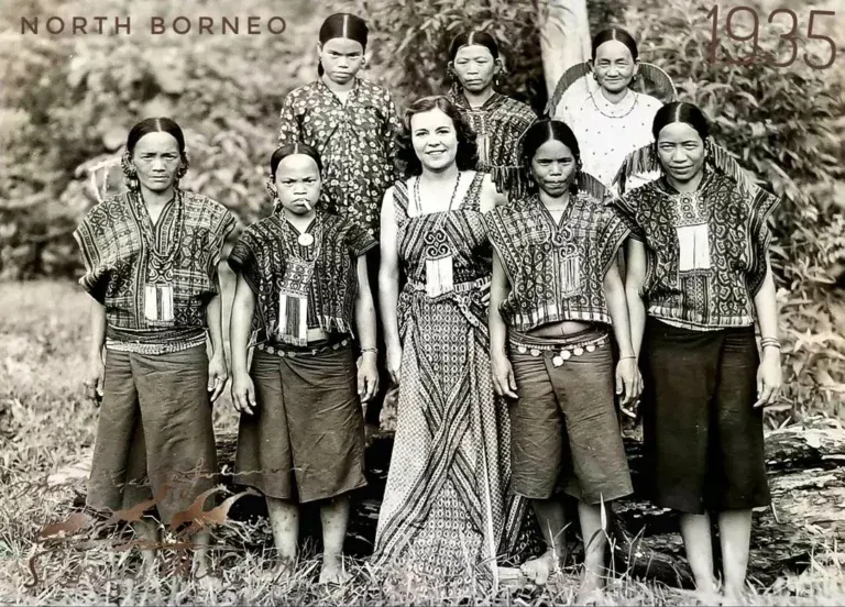 A group of nine people in traditional woven clothing, including a woman in a patterned dress, pose in North Borneo in 1935.