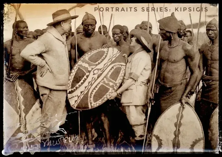 Two people in colonial-era attire interact with Kipsigis warriors carrying traditional patterned shields.