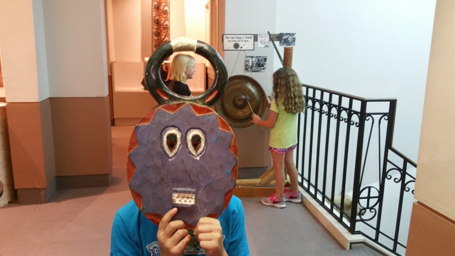 A child holds a purple, jagged-edged circular mask in front of their face near another child playing a large gong.