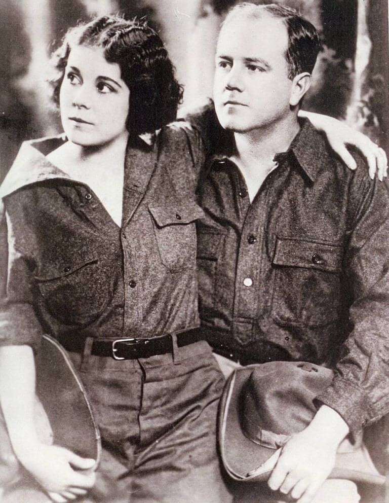 A black-and-white promotional portrait of two people wearing matching collared shirts and holding hats at their sides.