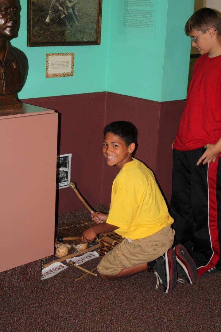 A person in a yellow shirt kneels on the floor, playing a percussion instrument in a museum exhibit.