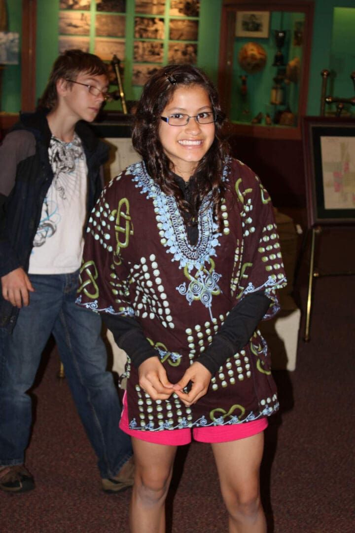 A smiling person wearing a patterned brown shirt and pink shorts stands in a museum, with a person walking behind them.
