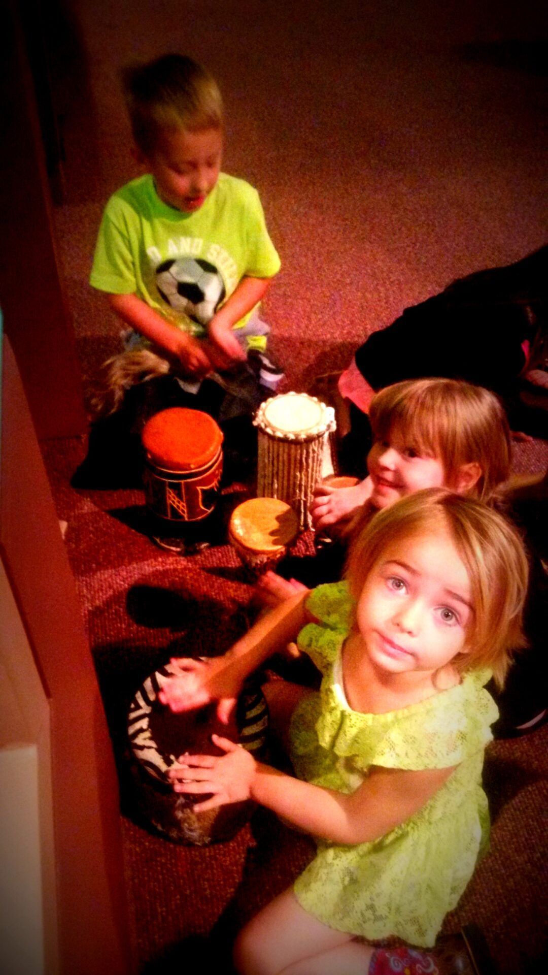 Three children in bright clothing sit on a carpeted floor, smiling while playing small drums.