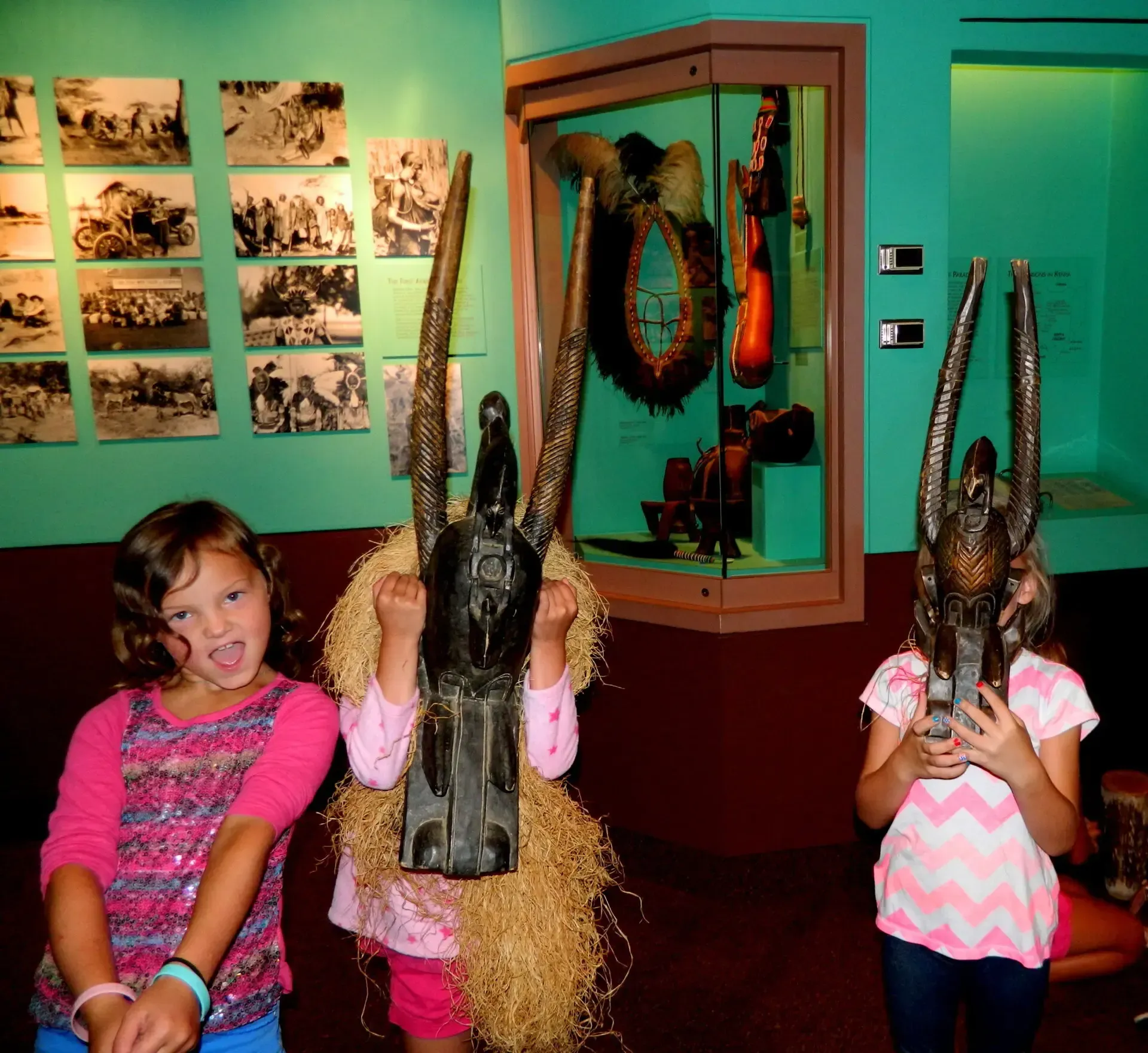 Three children in a museum display, two wearing elaborate carved wooden masks with tall horns and straw fringe.