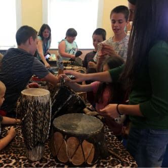 A group of people gathers around a table with decorative drums to learn music in a classroom setting.