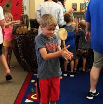 Children explore musical instruments in a classroom. A boy holds a wooden mallet in front of him with a playful expression.