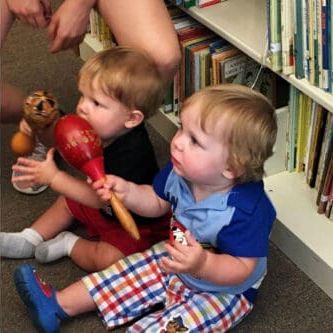 Two children sit on a floor in front of bookshelves, playing with a red maraca and a small figurine.