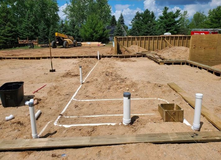 Construction site with marked foundation trenches, white pipes, and excavator in the background