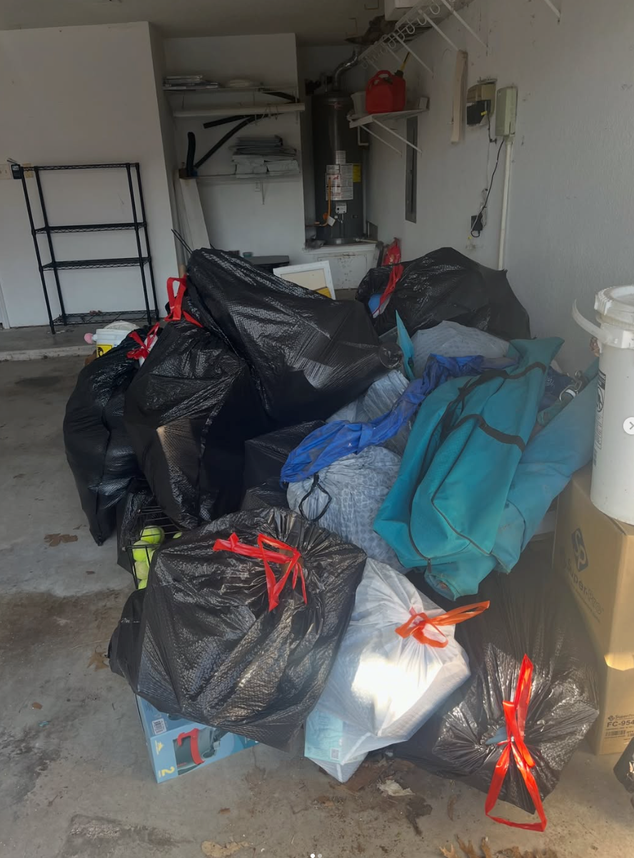 Garage cluttered with tied black trash bags, a blue bag, and shelves against the wall.