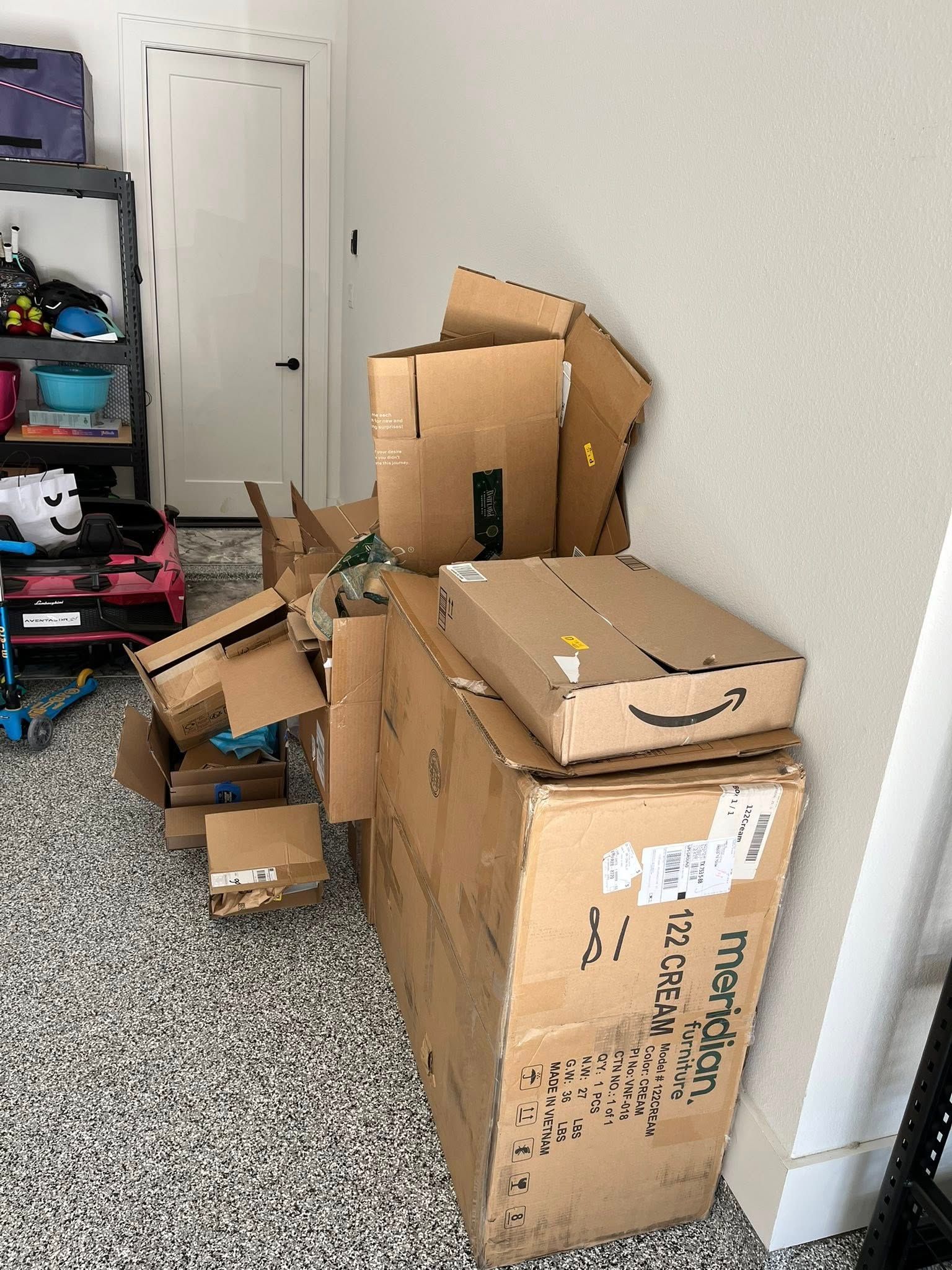 Cardboard boxes stacked along a wall in a small storage area beside a shelving unit and closed door.