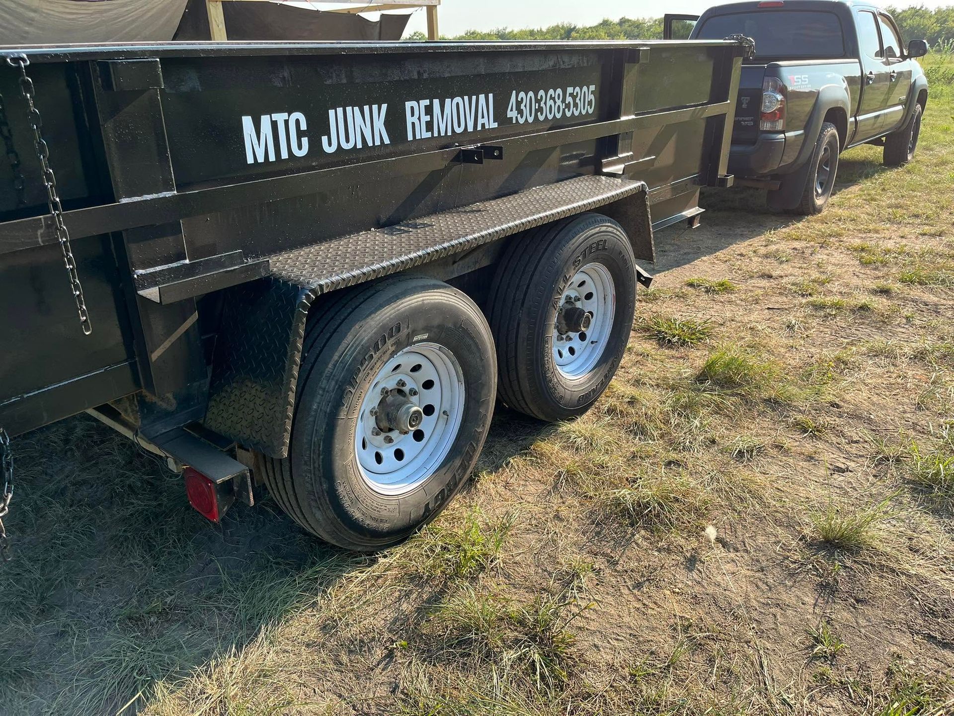 Black dump trailer hitched to a truck in a grassy field, with white lettering on the side.