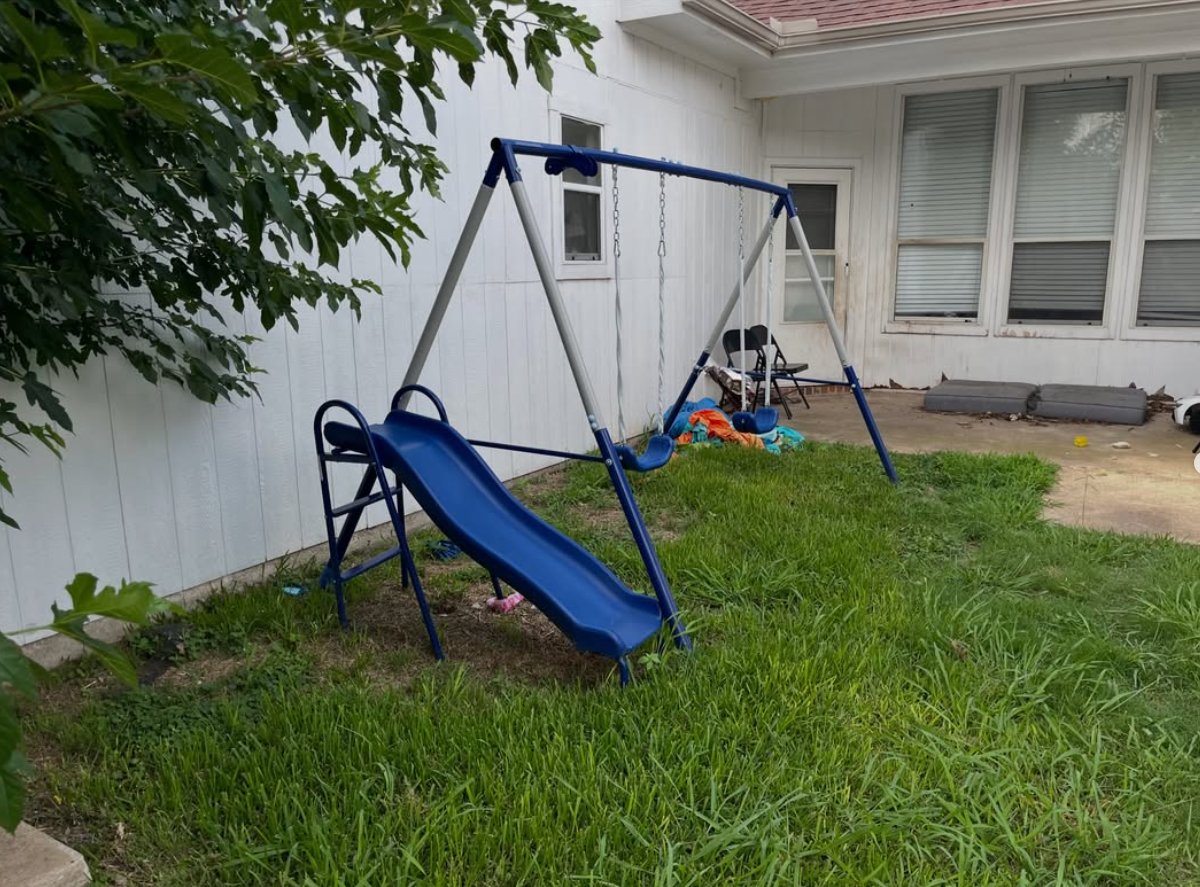 Blue backyard swing set and slide beside a white house, with scattered toys near the fence.