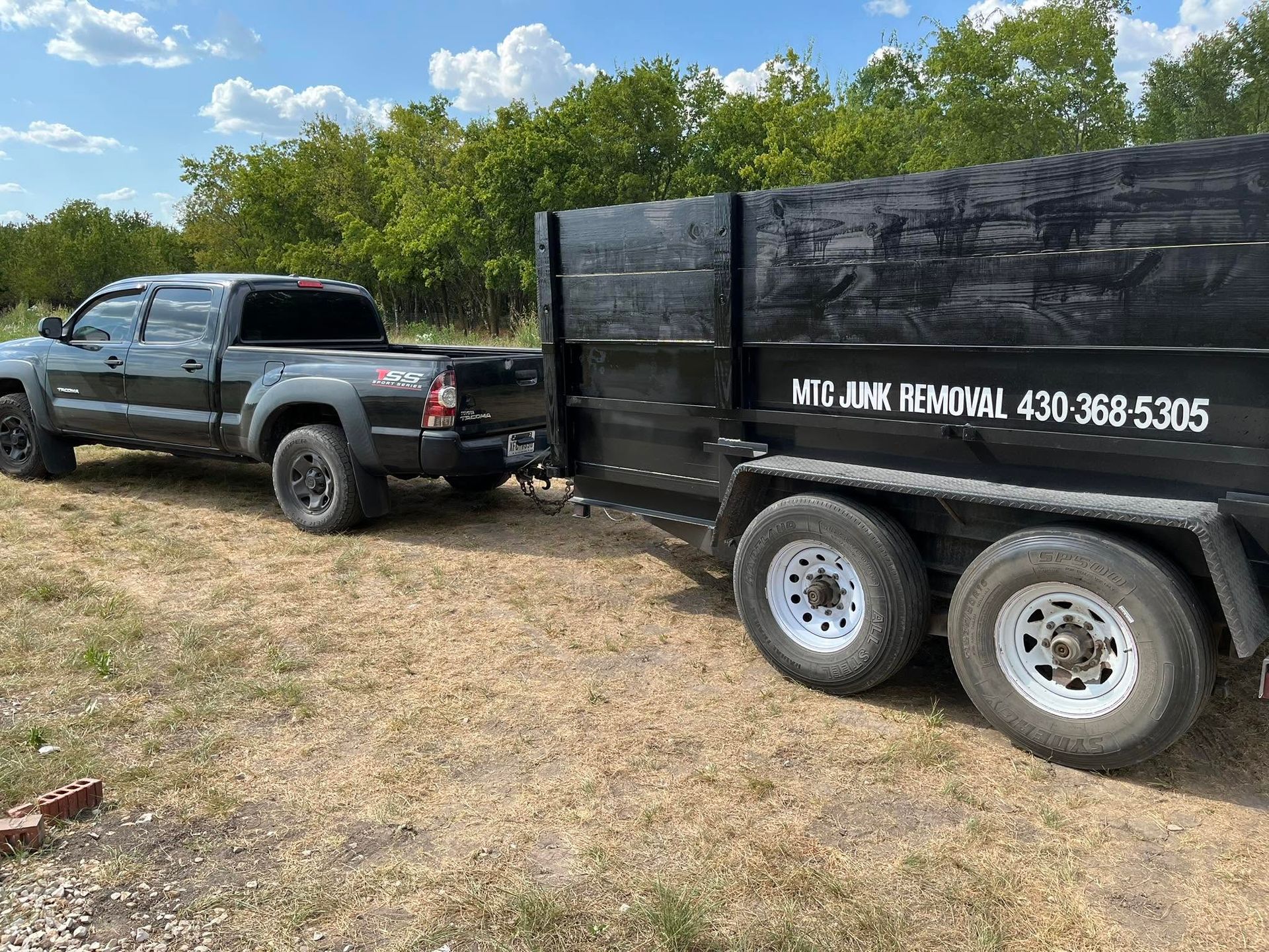 Gray pickup truck towing a black dump trailer in a grassy field, with trees and blue sky behind.