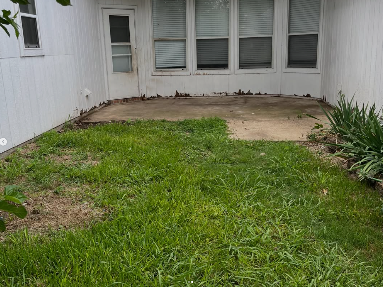 Small backyard patio with grass, a concrete pad, white house wall, and windows.