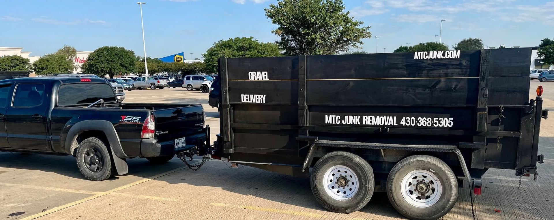 Black dump trailer with white lettering parked beside a dark pickup in a gravel lot under a blue sky.