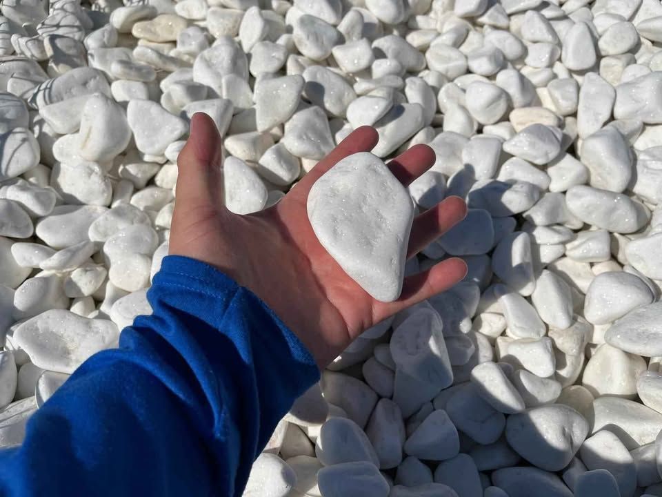 Hand holding a white stone over a bed of white pebbles, with a blue sleeve visible.