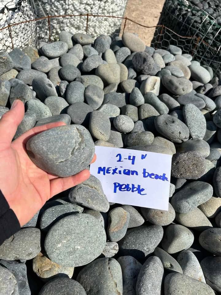 Hand holding a smooth gray stone among a pile of rocks with a “Mexican beach pebble” sign.
