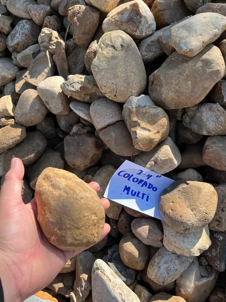 Hand holding a tan rock in front of a pile of stones, with a small handwritten label nearby.