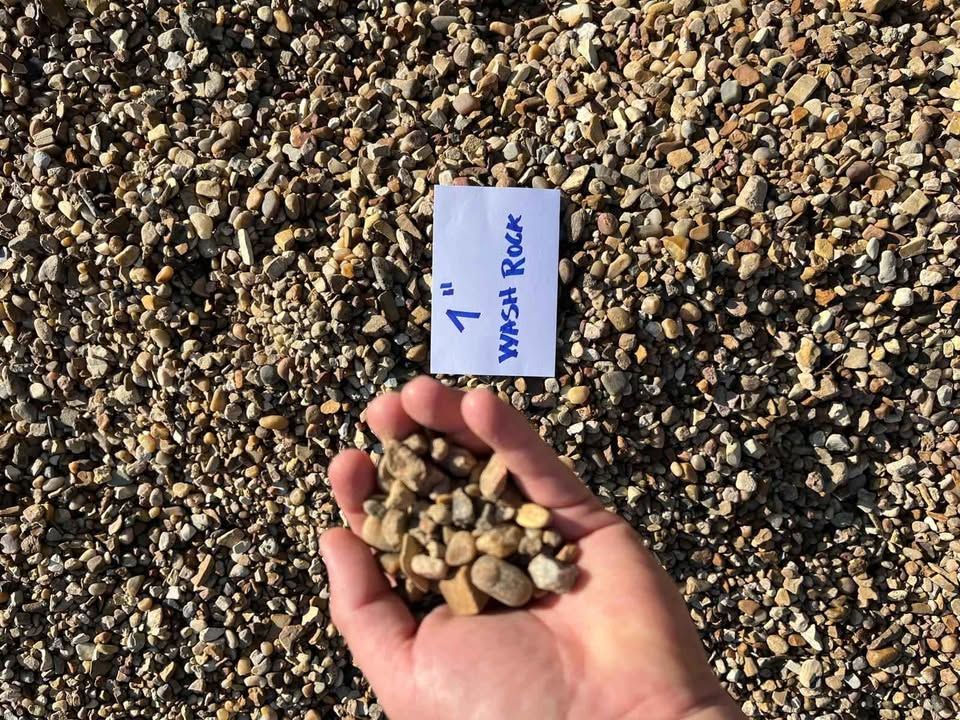 Hand holding small tan pebbles beside a labeled card on gravel, marked “Maui Rock.”