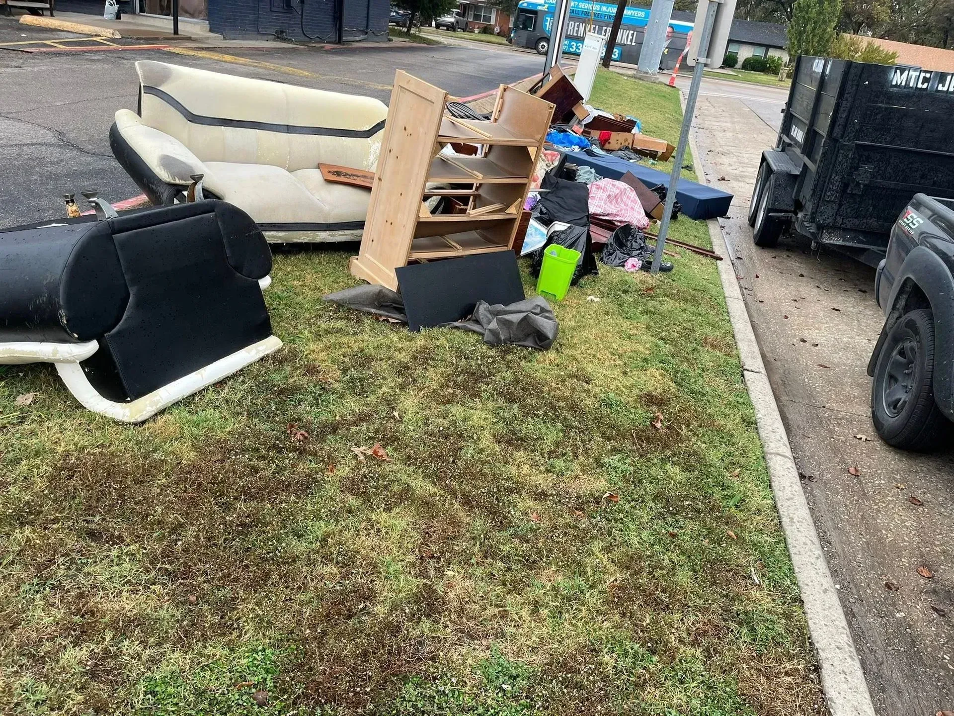 Curbside pile of discarded furniture, boxes, and bags on a grassy strip beside a road.