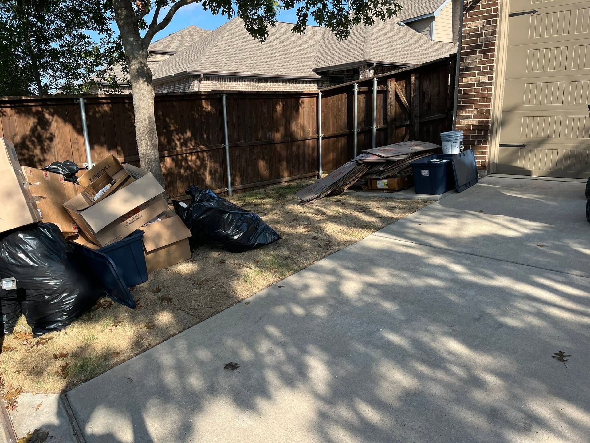 Driveway beside a garage with piles of trash bags, cardboard boxes, and debris stacked along a wooden fence
