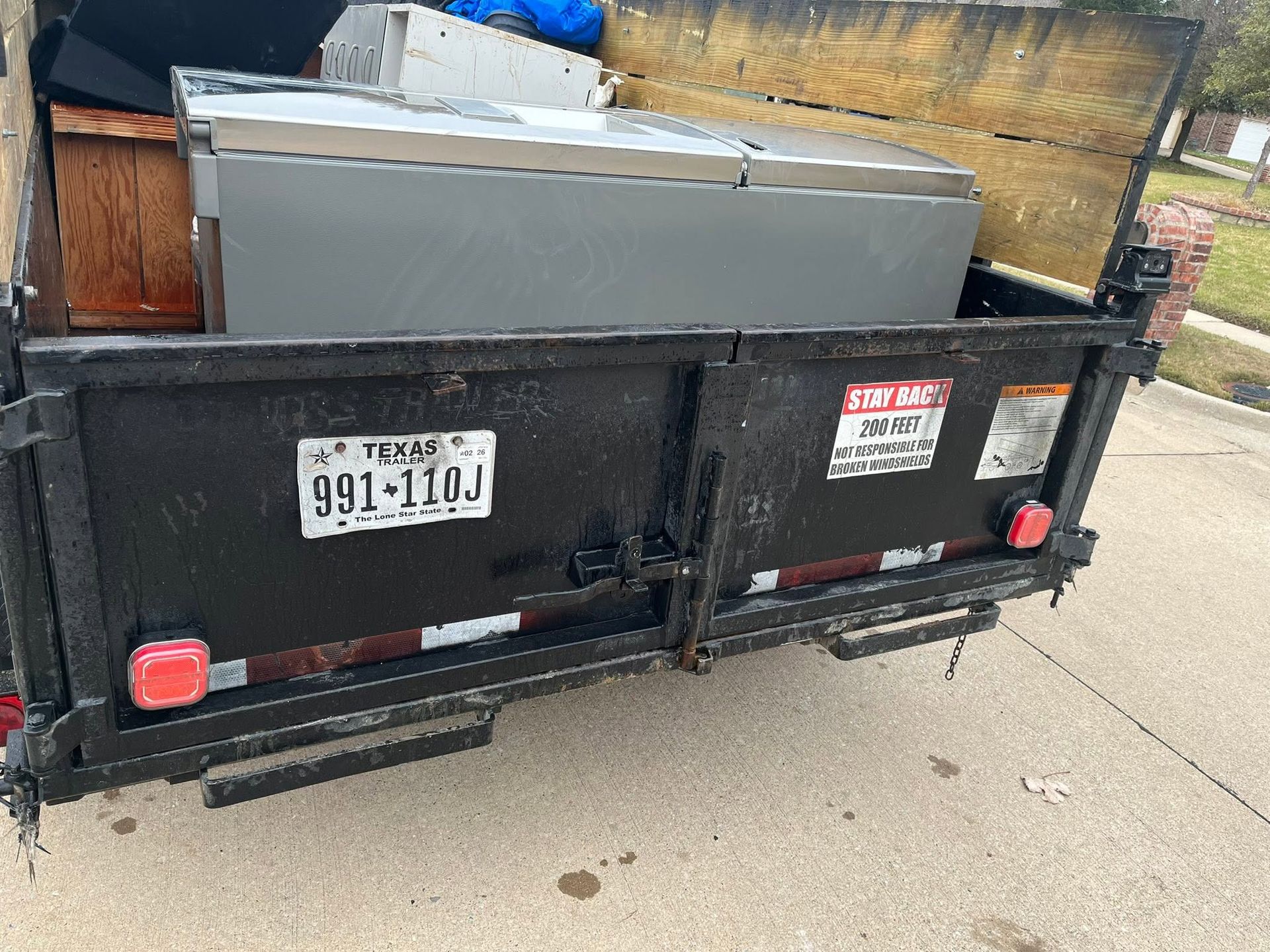 Black utility trailer loaded with a gray appliance and scrap wood on a driveway, with license plate visible