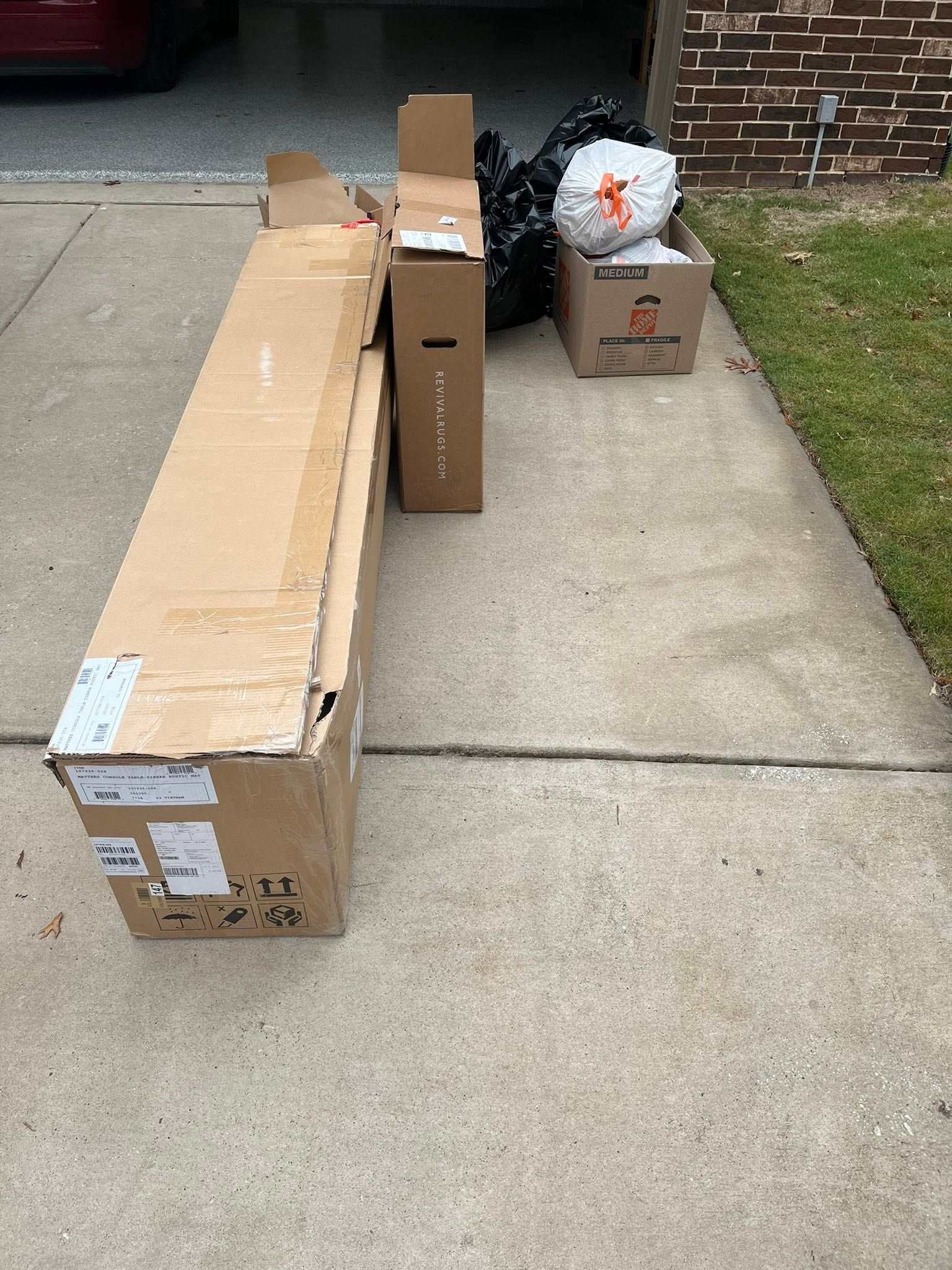 Cardboard boxes and a bag on a sidewalk beside a brick building and grass.