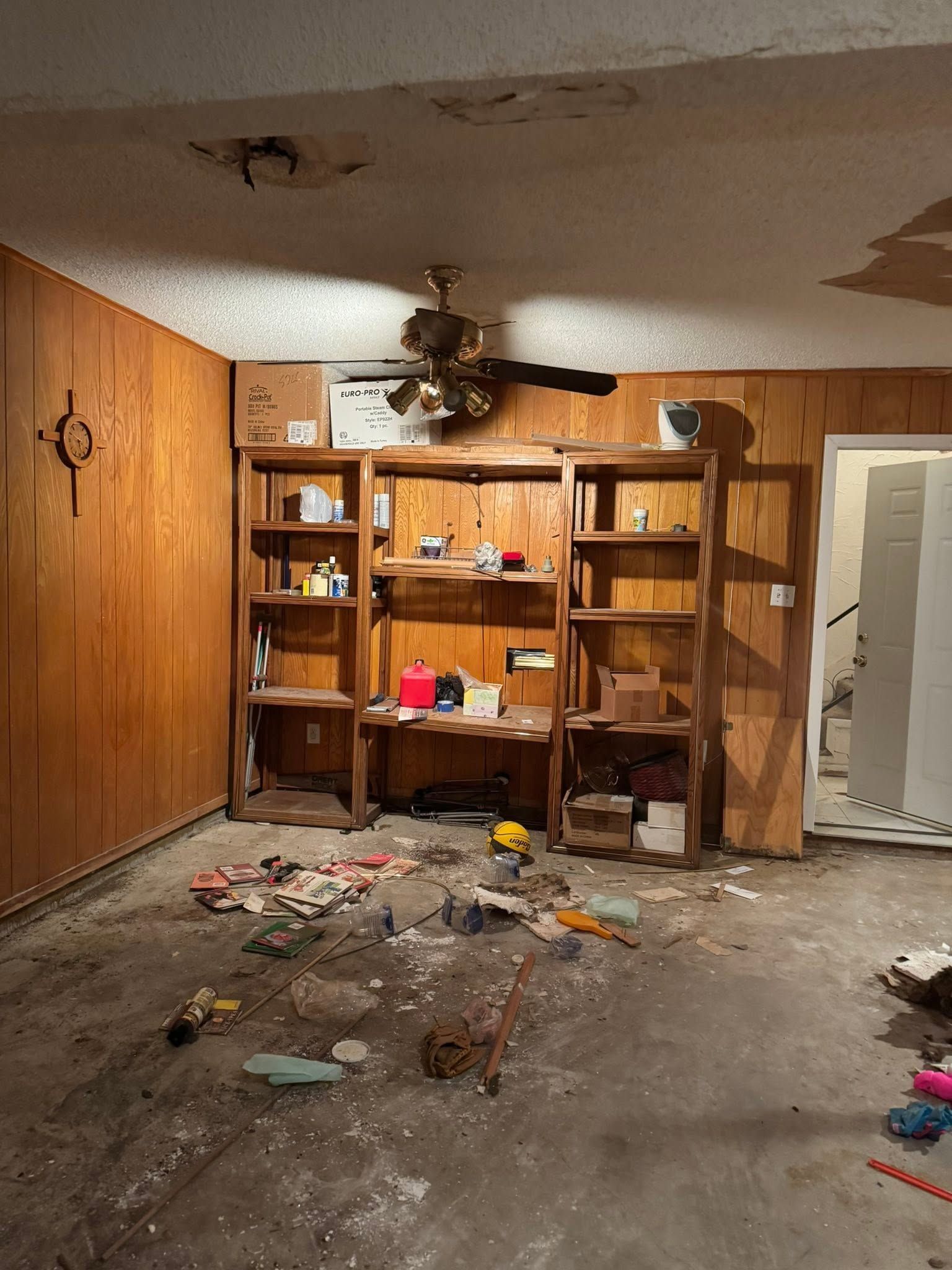 Damaged, messy room with broken ceiling, scattered debris, and a wooden shelving unit against the wall