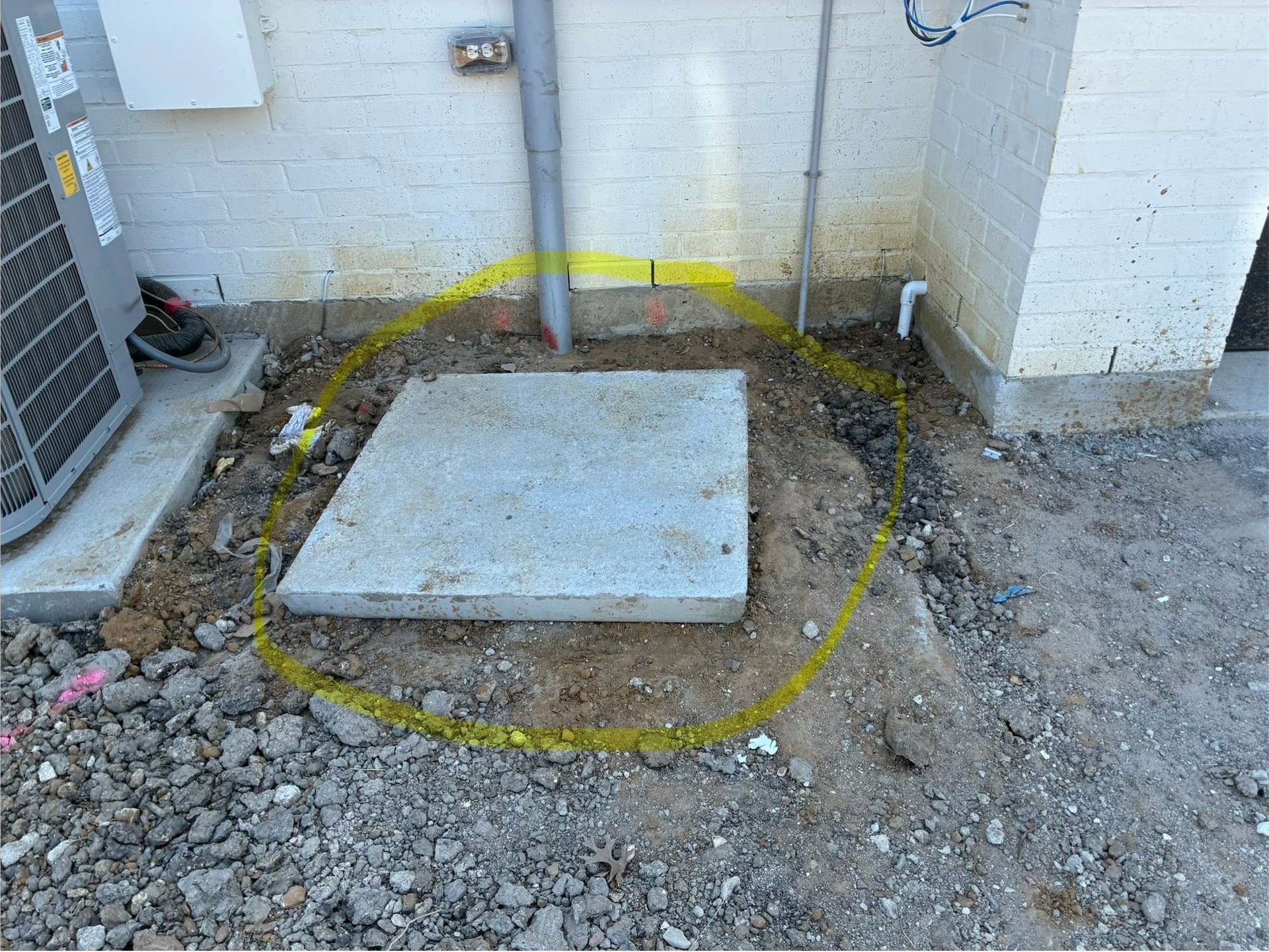 Concrete slab marked by yellow paint on gravel beside a brick wall and utility pipes.