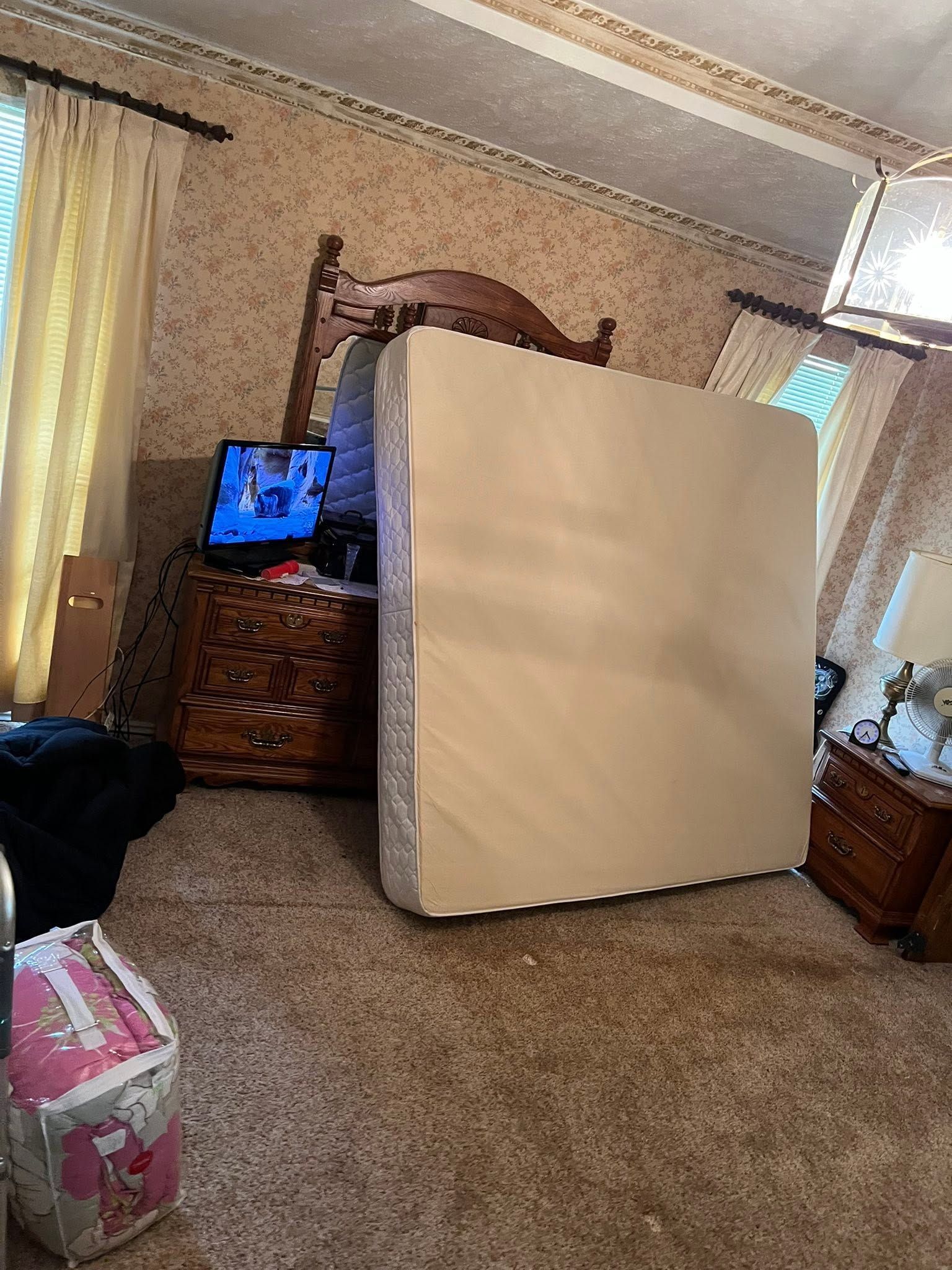 Mattress leaned against a dresser in a carpeted bedroom with windows and a TV.