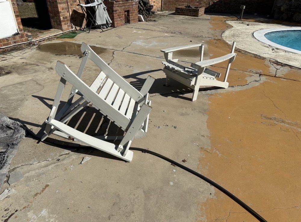 Collapsed white patio chairs on a cracked pool deck beside muddy floodwater.