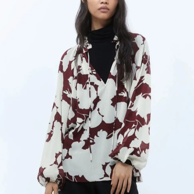 Mujer que lleva una blusa color crema con estampado floral granate sobre un cuello alto negro; contra un fondo blanco.