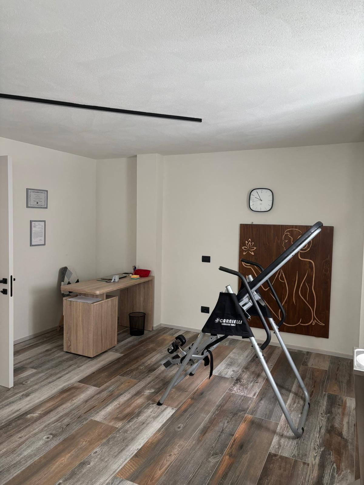 Stanza spoglia con pavimento effetto legno, piccola scrivania, telescopio montato su treppiede e orologio a parete.