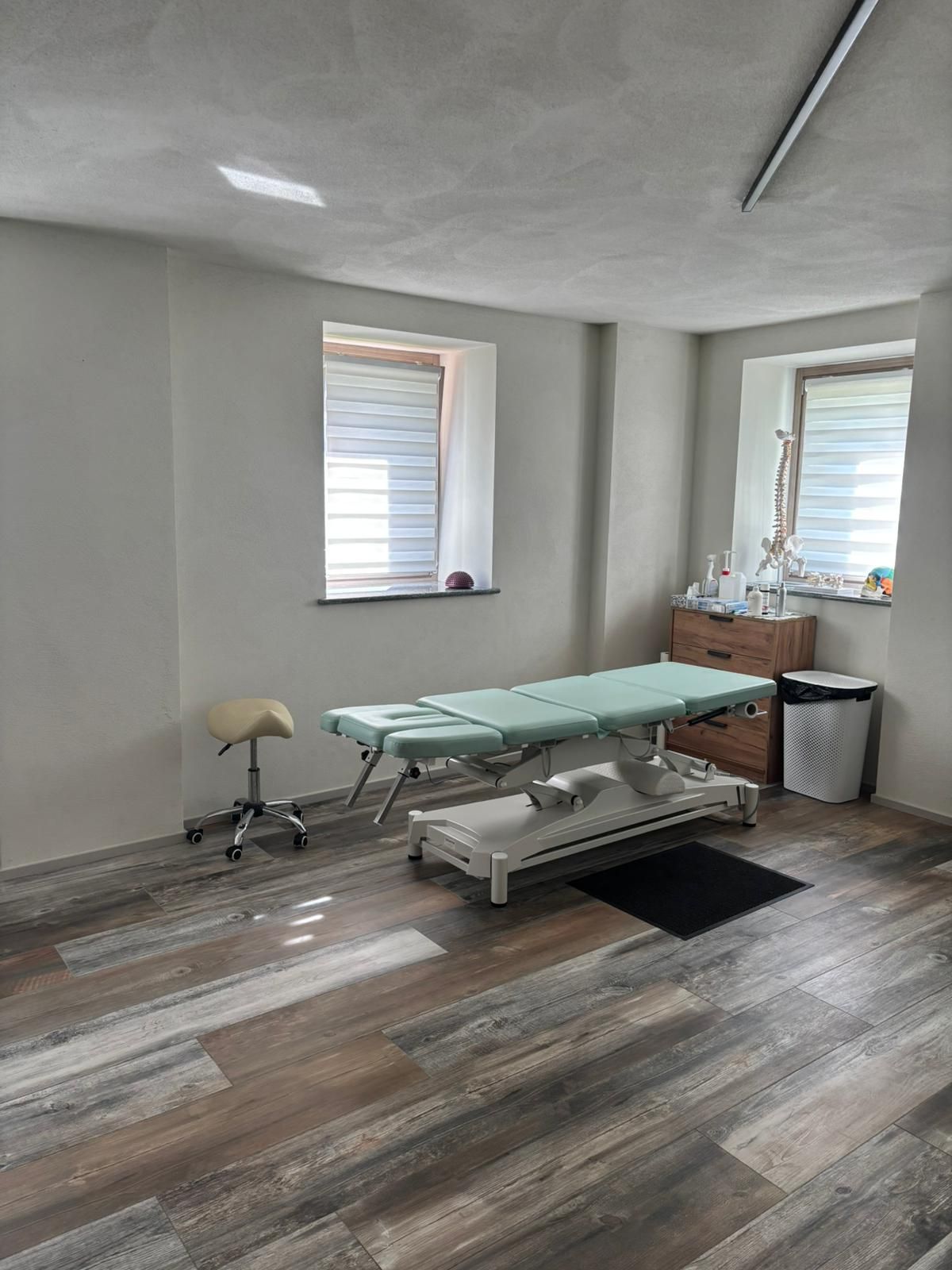Sala trattamenti vuota con lettino da massaggio, tende alle finestre e pavimento effetto legno.