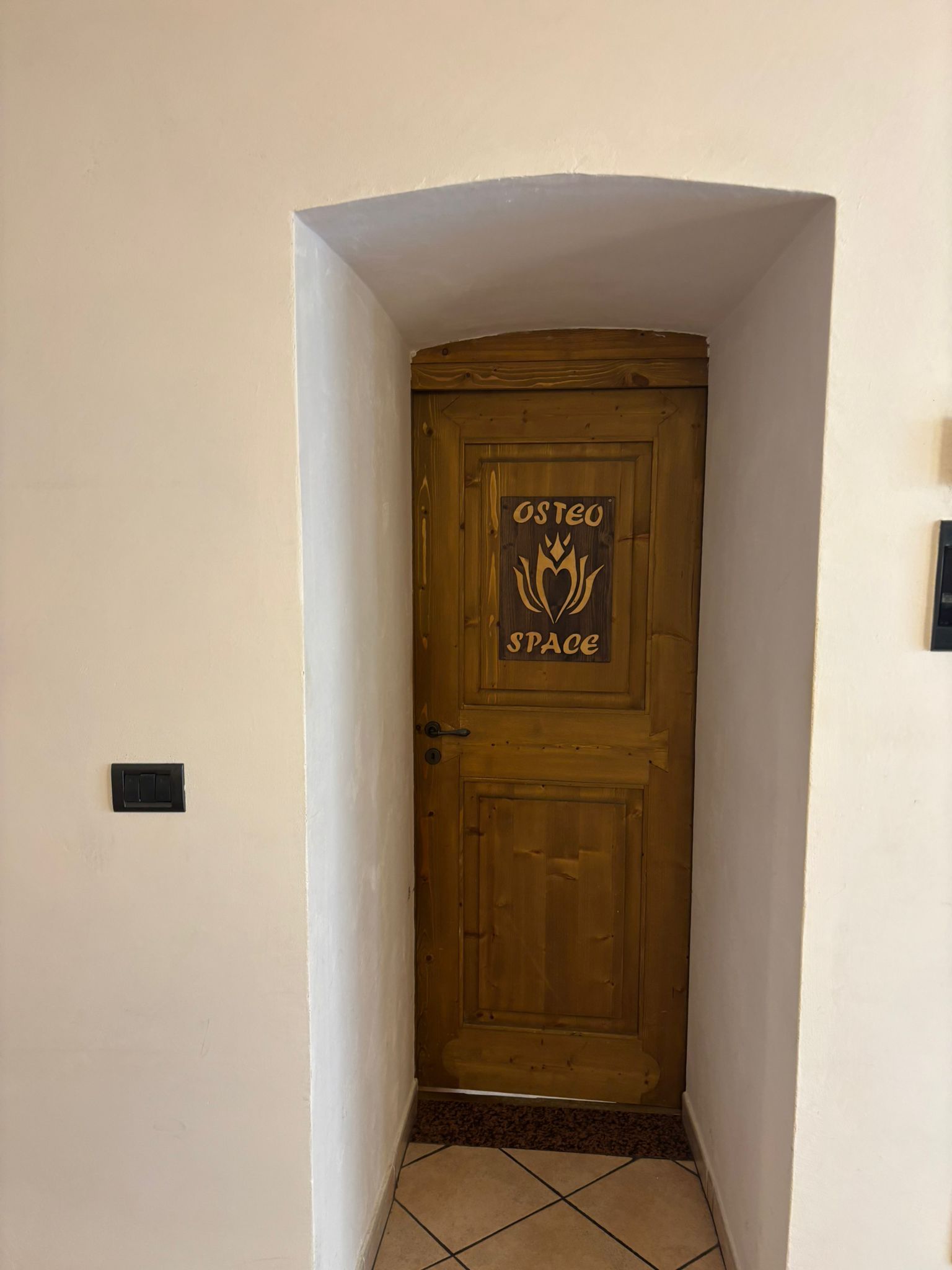 Nicchia stretta e bianca con porta in legno e targa decorativa, piccolo interruttore a parete nero sulla sinistra.