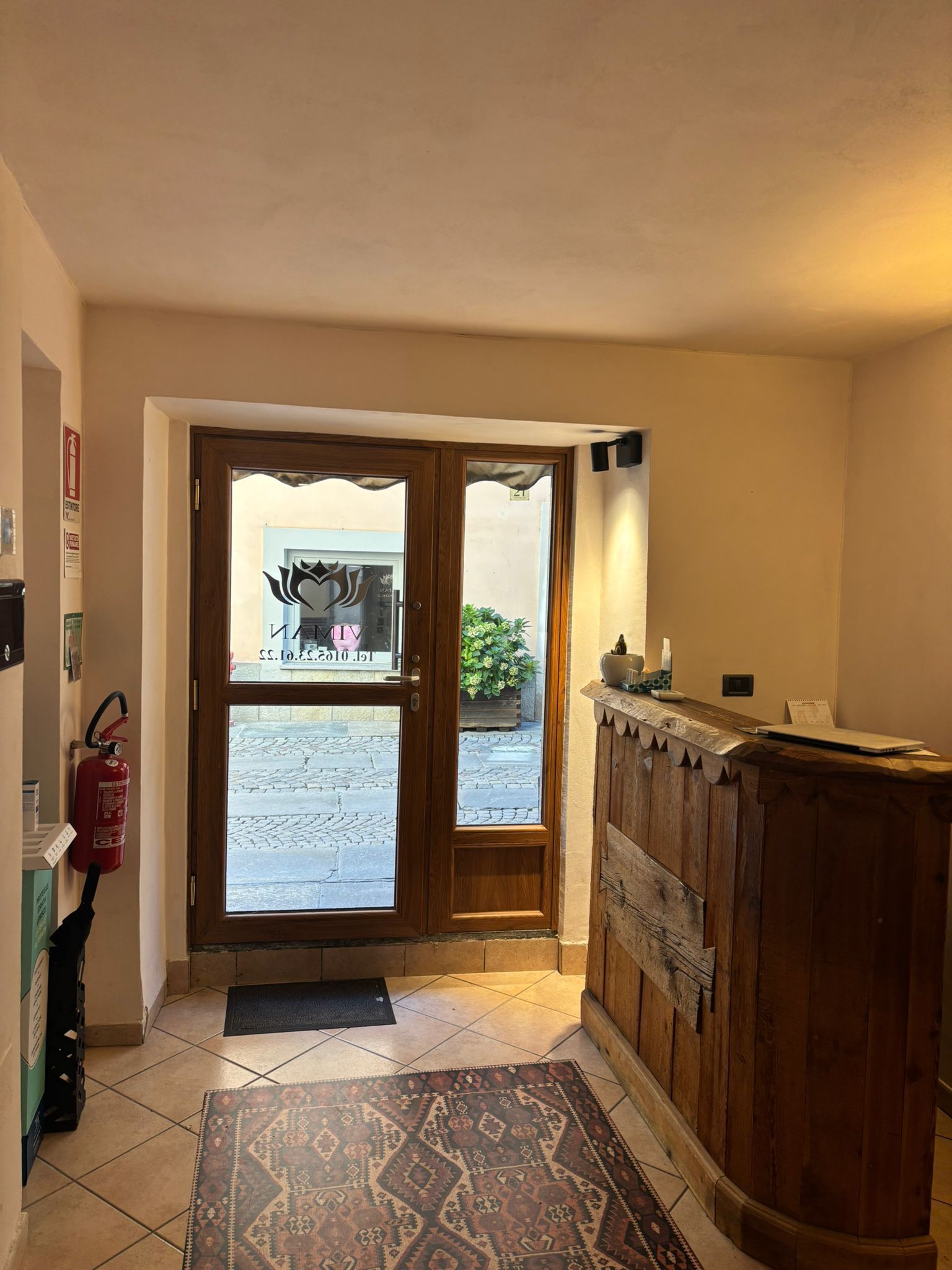 Atrio rivestito in legno con porte d'ingresso in vetro, un tappeto decorato e luce naturale proveniente dall'esterno.