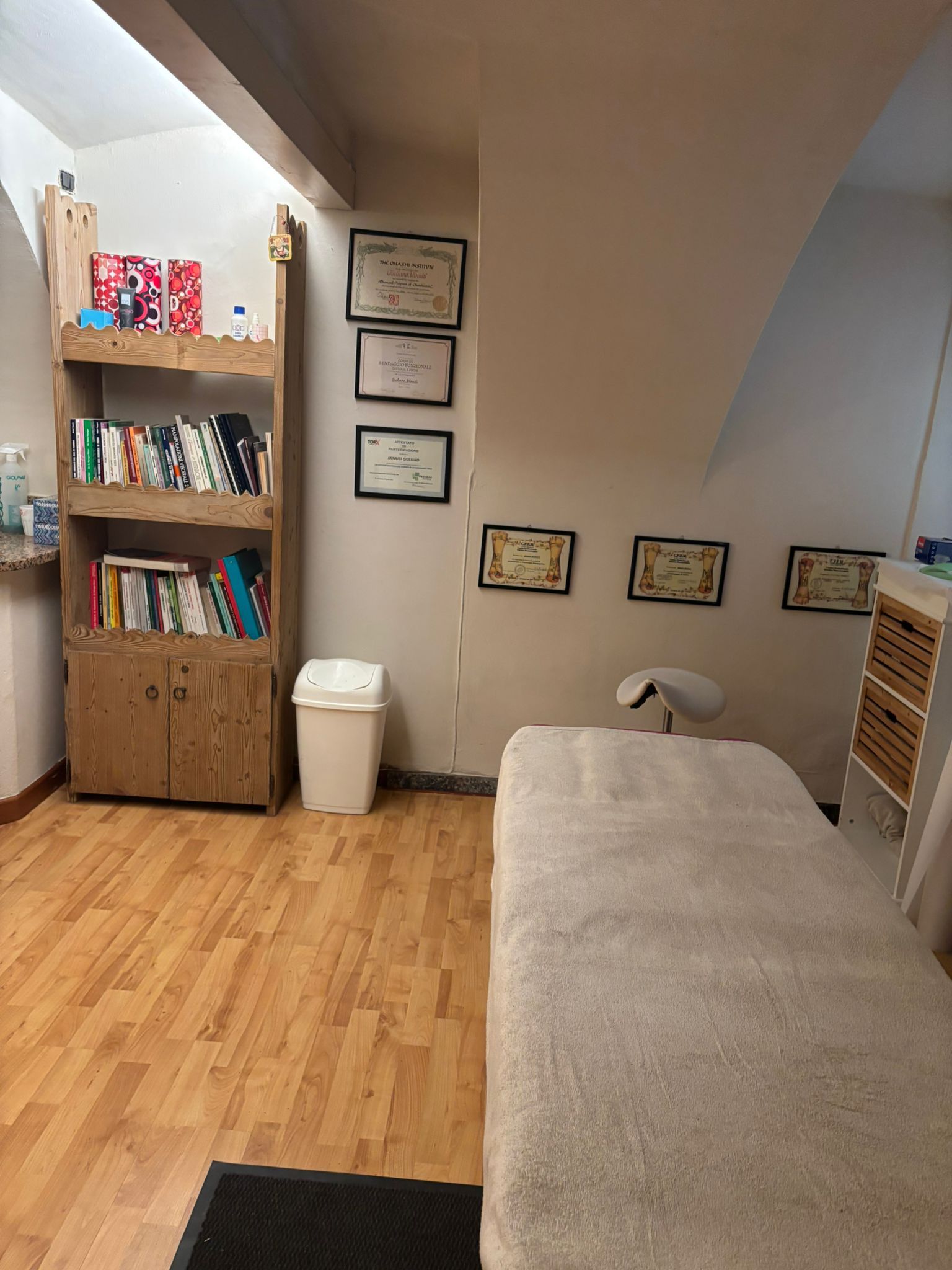 Mansarda con librerie, quadri incorniciati, un cestino bianco e un letto o divano letto con lenzuola grigie.