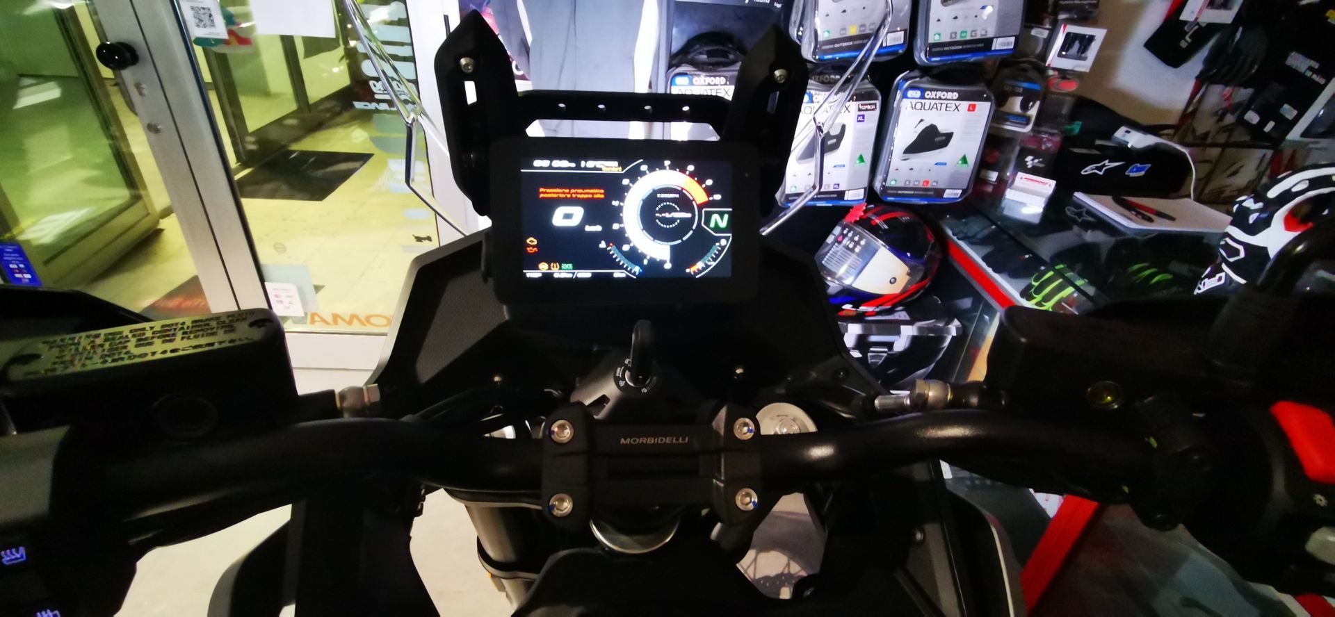 Cruscotto di una motocicletta con display digitale, manubrio e parabrezza. Sullo sfondo, l'interno di un'officina.