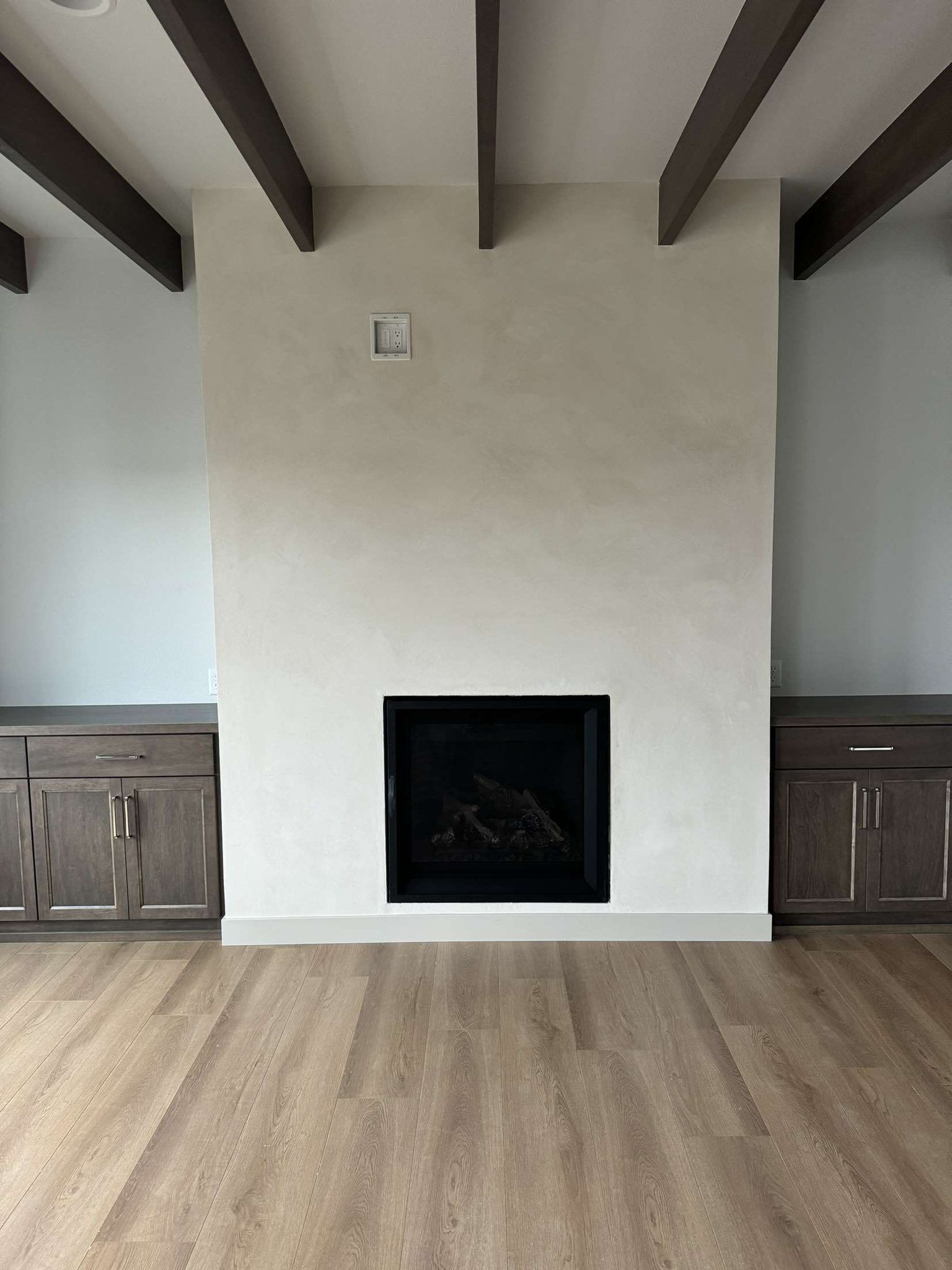 Fireplace with off-white wall, dark wood cabinets, and brown ceiling beams.