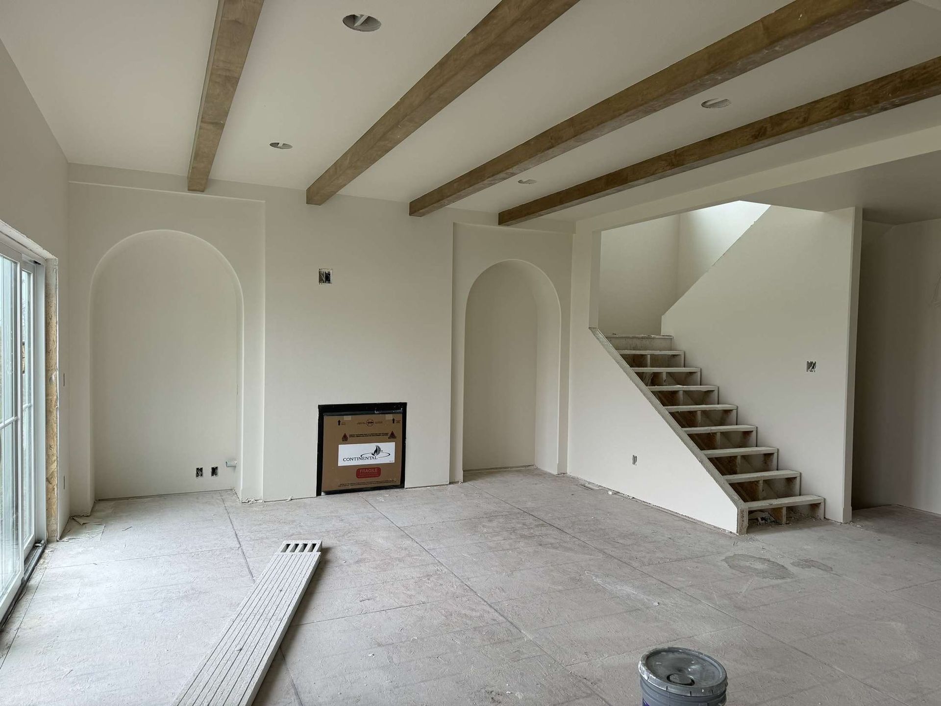 Unfinished interior with white walls, wooden beams, arched alcoves, and a staircase; concrete floor.